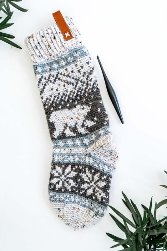 A knitted stocking with a bear pattern, next to a black crochet hook, on a white background. Green leaves frame the edges.