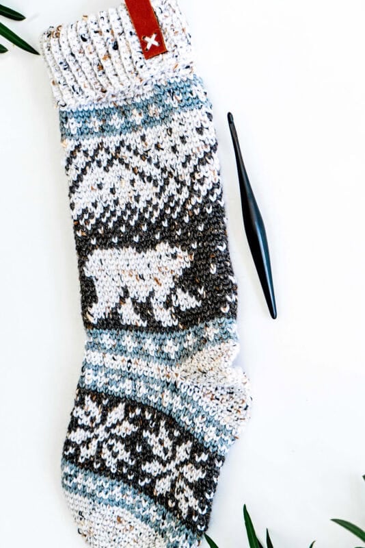 Knitted sock with a polar bear pattern, accompanied by a black crochet hook on a white background, surrounded by green leaves.