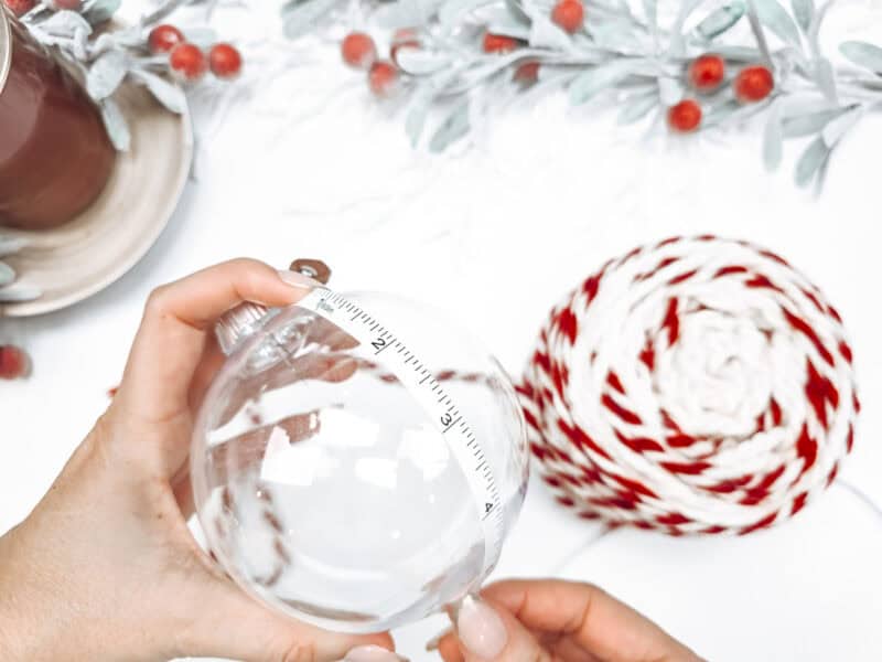 Hands measuring a clear Snowy Wheat Ornament with a measuring tape, next to red and white yarn and decorative red berries on a white surface—a perfect scene for crafting with our free crochet pattern.