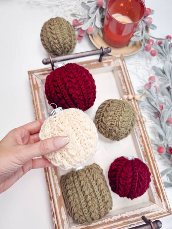 Hand holding a white knitted ornament above a tray with red and green knitted ornaments, next to a red candle on a decorated table.