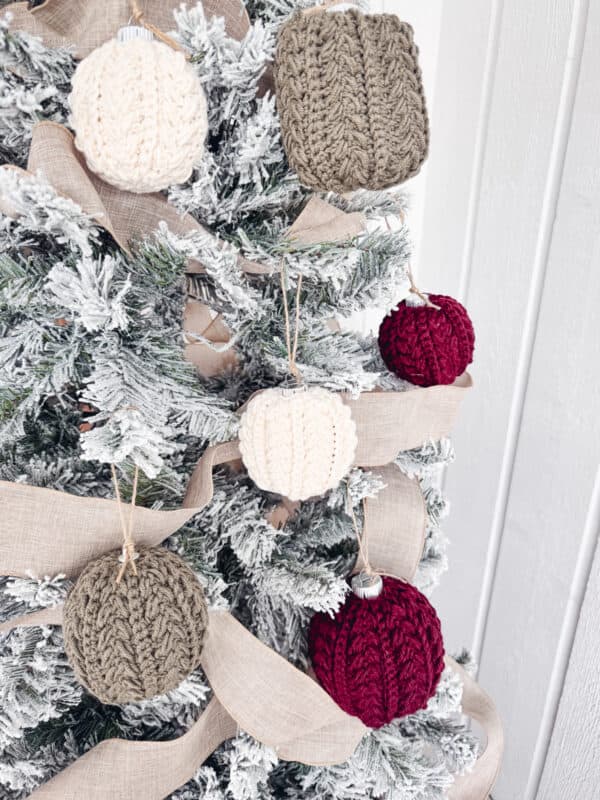 A snow-covered tree decorated with knitted ornaments in white, gray, and red, with beige ribbon wrapped around it.