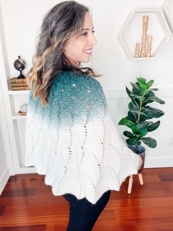 Woman wearing a green and white knit shawl stands beside a plant in a modern, decorated room.