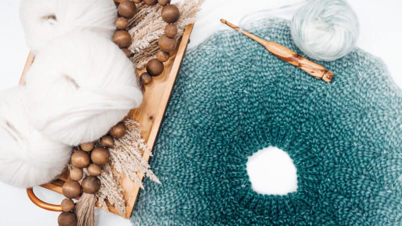 Crochet project in progress with teal and white yarn, wooden crochet hook, and decorative elements on a wooden tray.