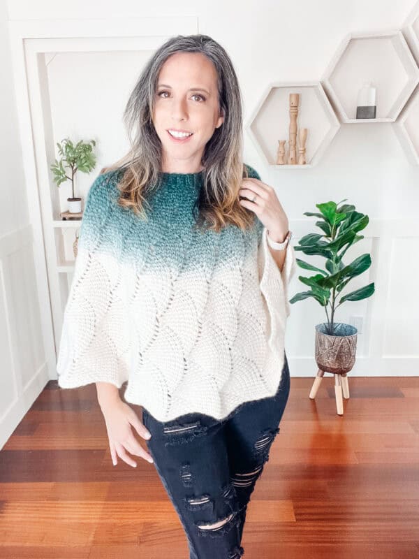 A person stands in a room with geometric wall shelves and a plant, wearing a green and white knitted poncho and ripped jeans.