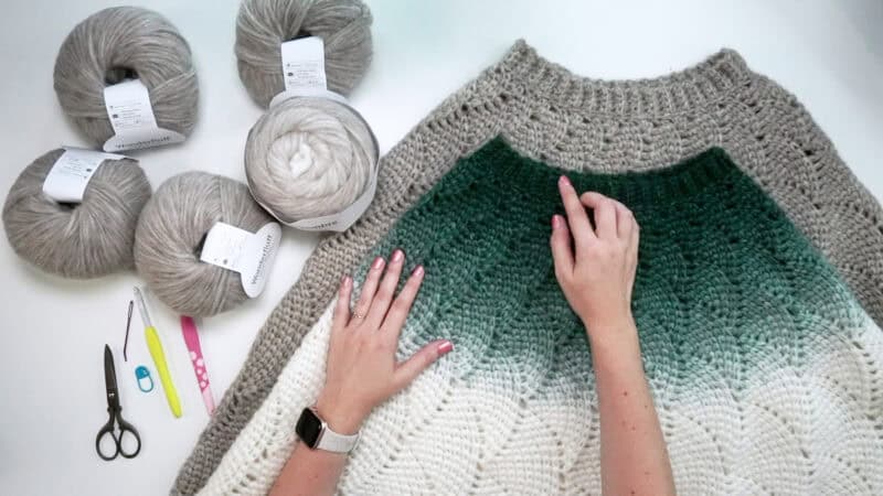 Hands working on a green to white gradient crochet piece, surrounded by gray yarn balls, scissors, needles, and a crochet hook.