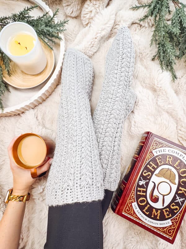 A person wearing cozy gray Snowdrift Crochet Slipper Socks, holding a mug beside a candle and a Sherlock Holmes book on a fluffy surface.