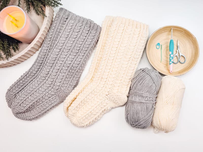 Gray and cream knitted socks, two balls of yarn, crochet hooks, and scissors arranged on a white background. A lit candle and greenery are partially visible.