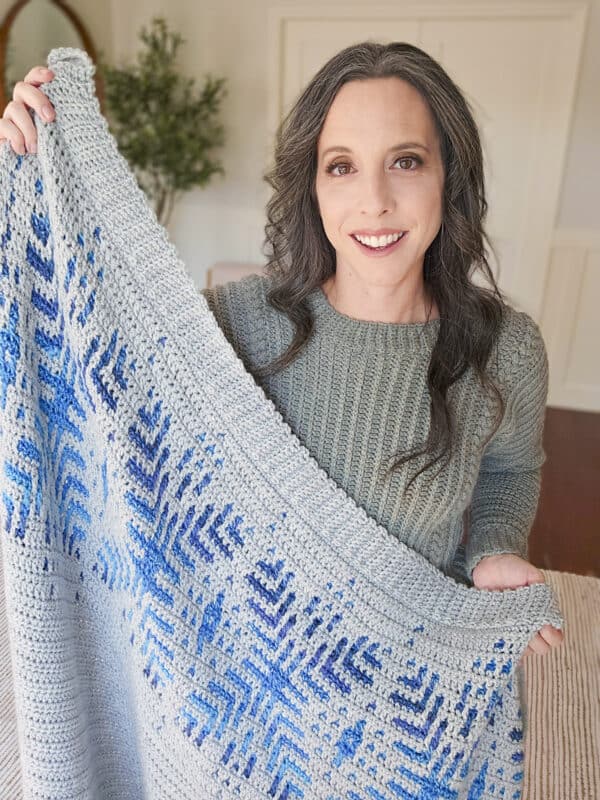 Woman holding a gray and blue knitted blanket with a geometric pattern, wearing a green sweater, smiling.