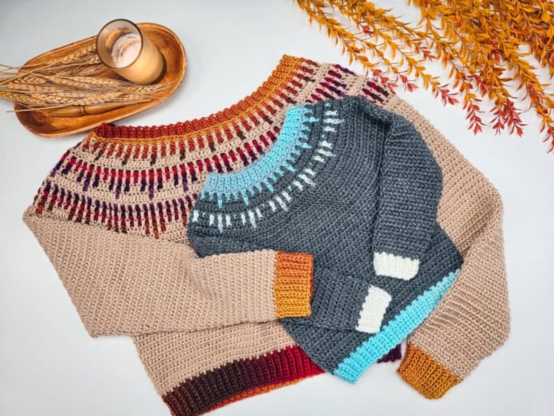 Two knitted sweaters with colorful patterns are laid on a surface. A decorative arrangement with a candle and wheat stalks is beside them.