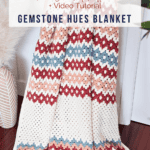 A beautifully draped crochet blanket with zig-zag patterns in vibrant gemstone hues of red, blue, and cream rests on a ladder. Text overlay reads: "Gemstone Hues Crochet Blanket Free Pattern + Video Tutorial, www.brianakdesigns.com.