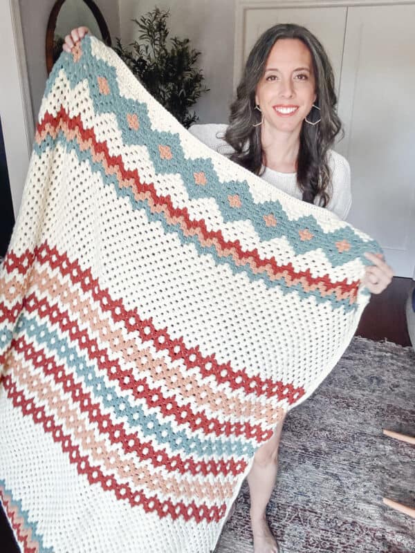 A person holding a crocheted blanket with a geometric pattern in gemstone hues of blue, red, and white.