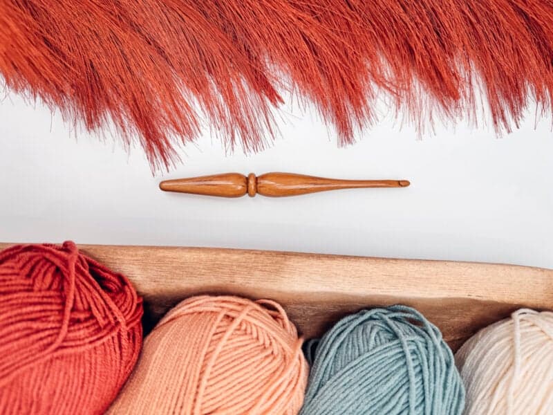 A crochet hook lies on a white background with a fringe of reddish yarn above, hinting at the creation of a Gemstone Hues Crochet Blanket. Below, a wooden tray showcases yarn balls in vibrant red, peach, blue, and white. Access the free pattern to bring this colorful vision to life.