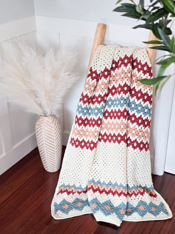 A vibrant crochet blanket in gemstone hues with intricate geometric patterns is draped over a wooden ladder. A vase filled with pampas grass sits gracefully on the floor to the left, adding a touch of natural elegance.