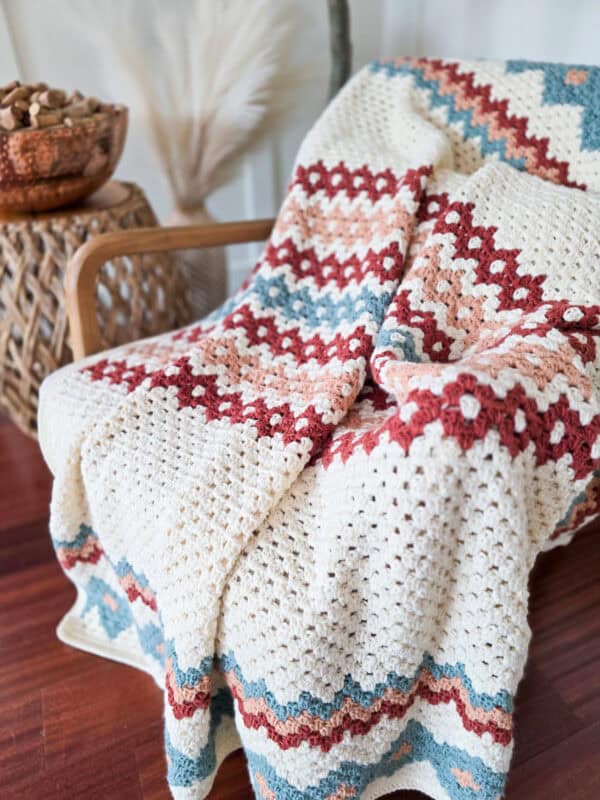 A crocheted blanket with gemstone hues in red, blue, peach, and white forms a zigzag pattern as it drapes elegantly over a wooden chair beside a woven side table.