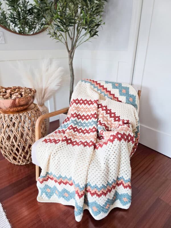 A chair draped with a crochet blanket in gemstone hues of red, blue, and cream. Nearby sits a woven basket and a tall plant in the corner against wood flooring.