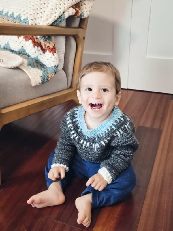 Smiling toddler in a gray and blue sweater sits on a wooden floor beside a chair with a crochet blanket.