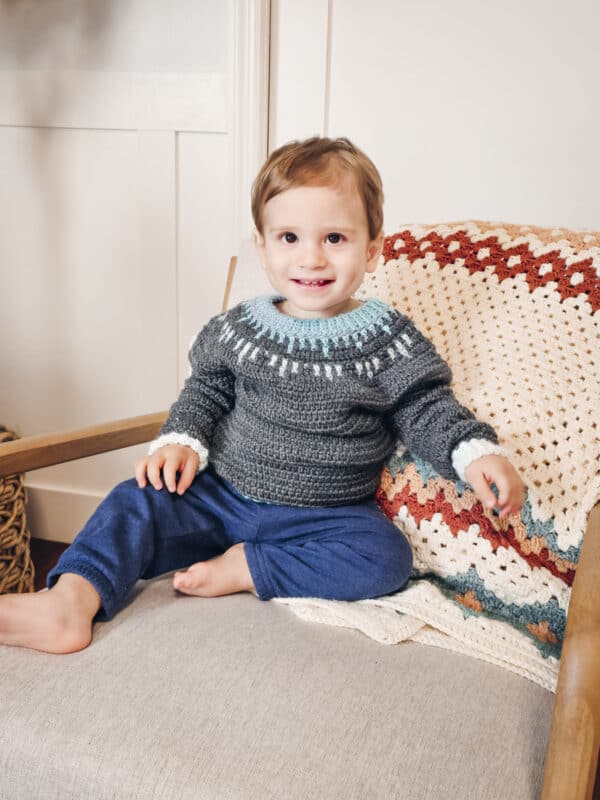 A toddler wearing a gray sweater with a blue and white pattern sits on a beige chair with a crocheted blanket, smiling.