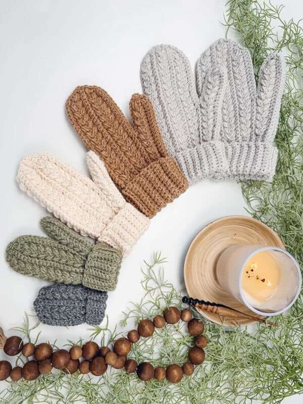 Four pairs of knitted mittens in various colors are arranged on a white surface next to a lit candle on a wooden coaster, surrounded by a string of wooden beads and green foliage.