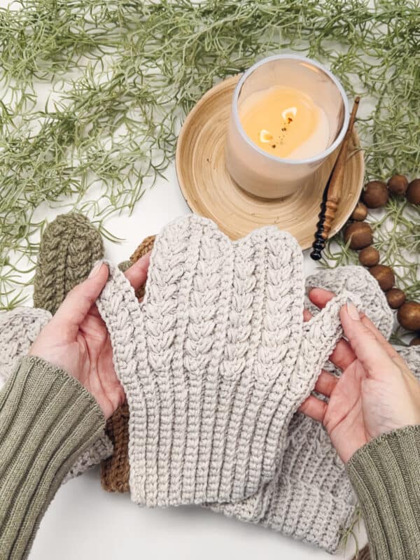 Hands holding a knitted beige mitten over a table with a lit candle, greenery, and other knitwear.