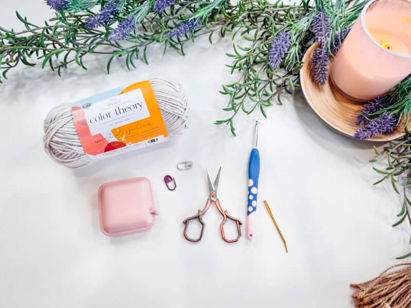 Yarn, crochet tools, scissors, stitch markers, and a candle on a white surface with sprigs of lavender.