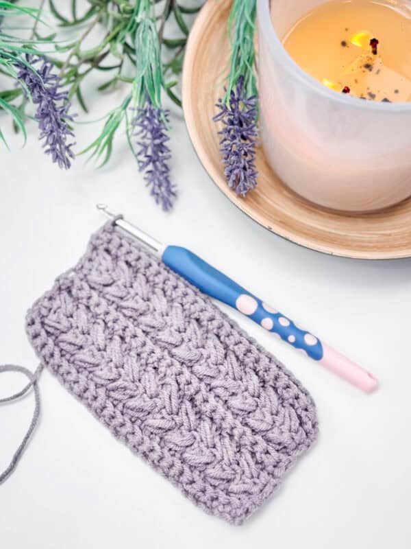 A small crocheted lavender-colored piece with a crochet hook lies next to a lit candle and sprigs of lavender on a white surface.