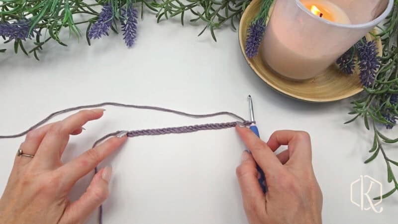 Hands crocheting a purple chain with a blue crochet hook, surrounded by lavender sprigs and a lit candle on a wooden plate.