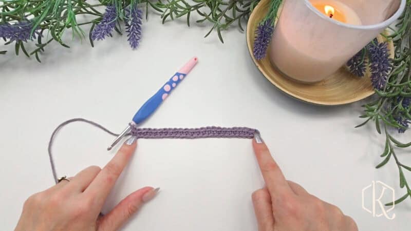 Hands pointing at a purple crochet piece next to a blue and pink crochet hook on a white surface; a lit candle and lavender sprigs are in the background.