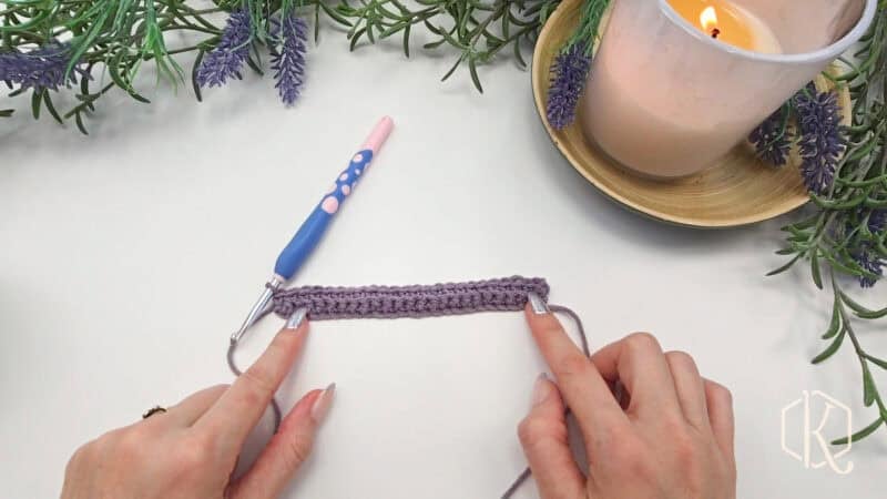 Hands holding a partially crocheted purple piece with a blue crochet hook nearby. Lavender sprigs and a lit candle are in the background.