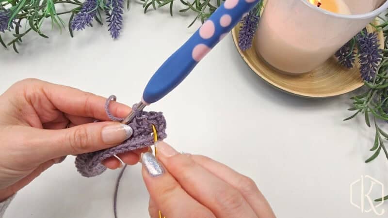 Hands crocheting with a blue and pink hook and a yellow needle, working on purple yarn. Lavender sprigs and a lit candle in the background.