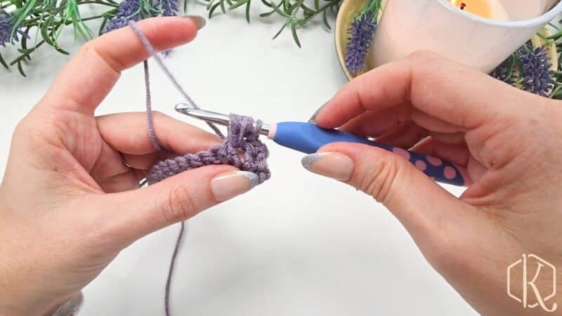 Hands crocheting with a purple yarn and a crochet hook, in front of lavender plants and a lit candle.