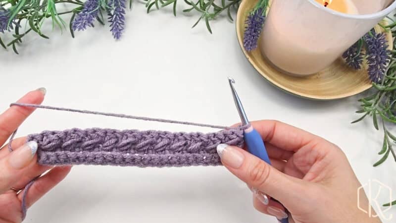 Hands crocheting with purple yarn and a blue-handled hook, surrounded by lavender sprigs and a lit candle.