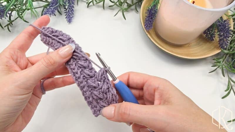 Hands crocheting with a purple yarn and blue-handled hook. Lavender sprigs and a lit candle in the background.