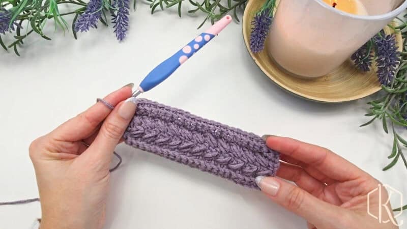 Hands holding a purple crocheted piece with a blue crochet hook nearby, surrounded by lavender sprigs and a lit candle.