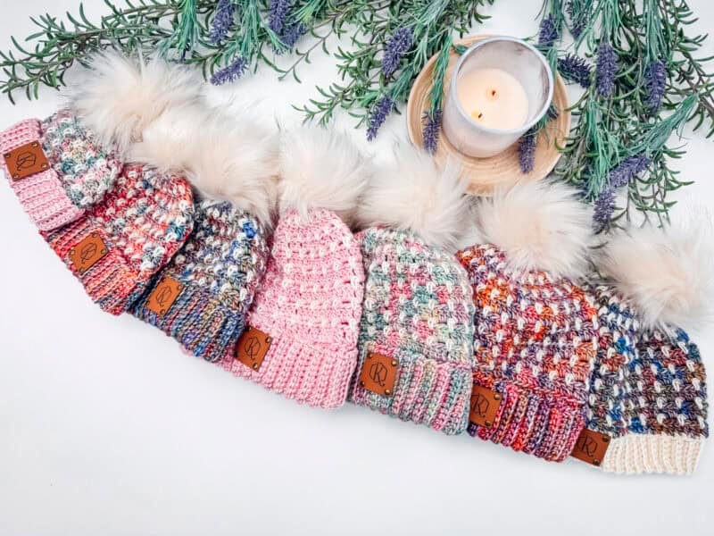 Six knitted beanies with pom-poms arranged in a row, each with different multicolored patterns. A lit candle and lavender sprigs are placed above them.