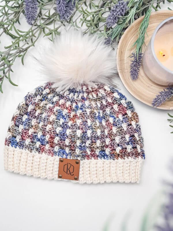 A multicolored knitted beanie with a fluffy pom-pom sits on a white surface, surrounded by lavender sprigs and a lit candle on a wooden tray.