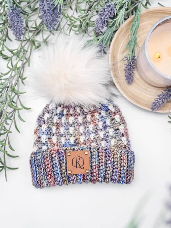 Multicolored knit beanie with a pom-pom on top, labeled with a leather patch. Surrounded by lavender sprigs and a lit candle on a wooden tray.