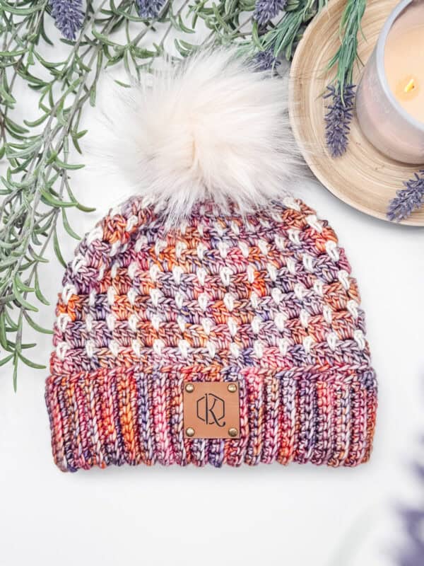 Crocheted multicolored beanie with a pom-pom, featuring purple, orange, and white patterns, and a leather patch on a white surface with greenery and a lit candle nearby.