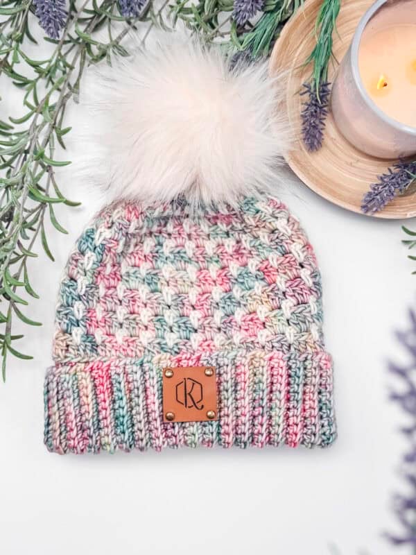 A multicolored knit beanie with a fluffy pom-pom on top, featuring a leather patch on the brim, surrounded by lavender sprigs.