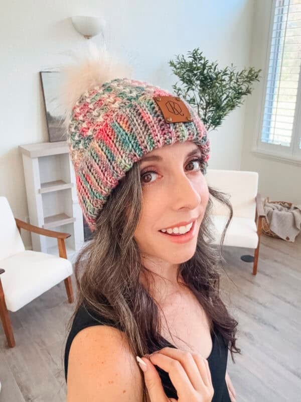 Person wearing a colorful knit beanie with a pom-pom, smiling indoors. Two chairs and a small shelf with plants are in the background.