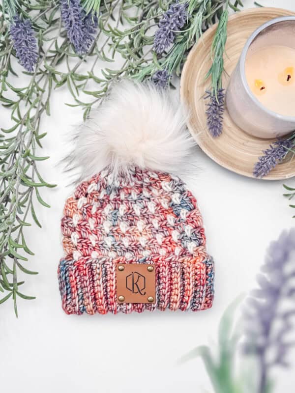 A multicolored knit hat with a fluffy pom-pom is surrounded by lavender sprigs. A lit candle on a wooden tray is nearby.