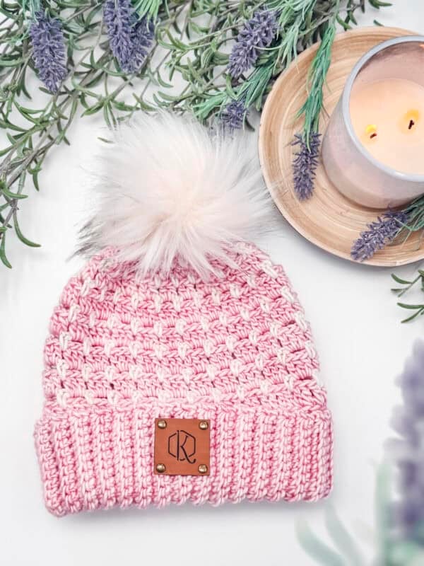 A pink knitted beanie with a fluffy pom-pom and a leather patch lies on a white surface, surrounded by lavender sprigs and a lit candle on a wooden tray.