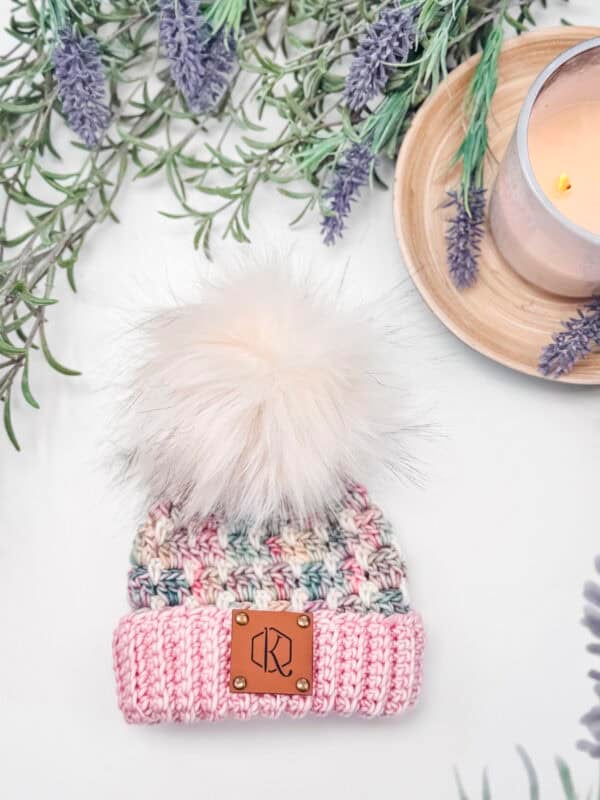 Knitted beanie with a fluffy pom-pom, surrounded by lavender sprigs and a lit candle on a wooden tray.