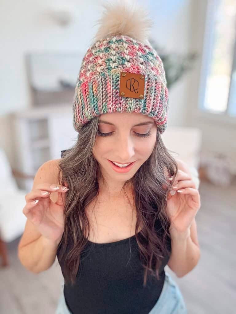 In a bright room, a person dons a black top and light denim pants, accentuated by a vibrant chunky crochet hat with a pom-pom, crafted using the block stitch pattern.