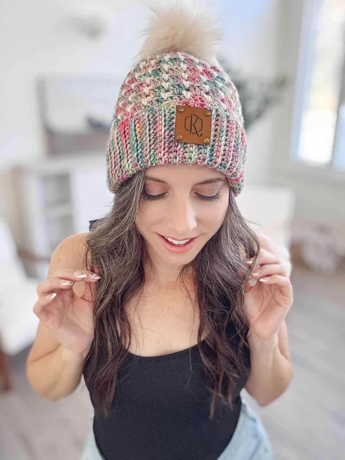 In a bright room, a person dons a black top and light denim pants, accentuated by a vibrant chunky crochet hat with a pom-pom, crafted using the block stitch pattern.