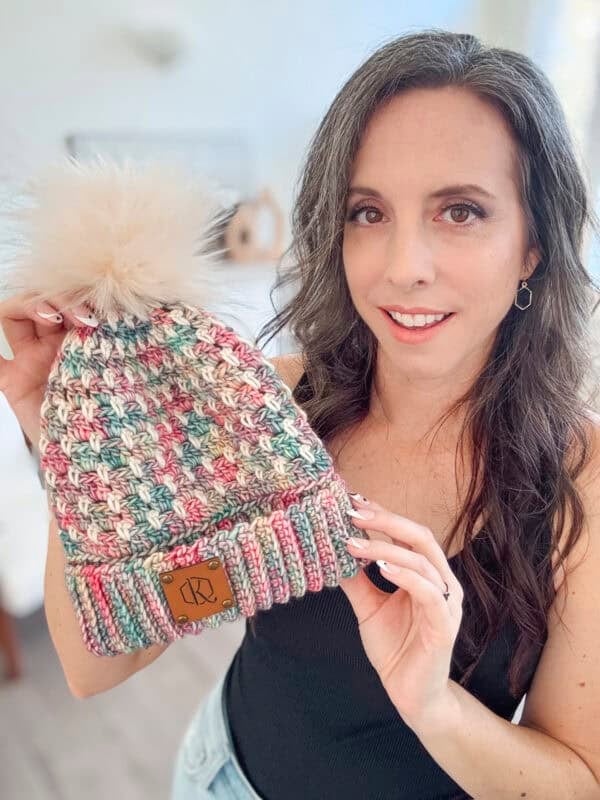 Person holding a multicolored knit beanie with a fluffy pom-pom, smiling at the camera.