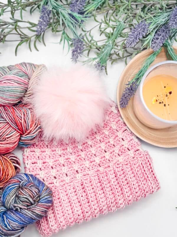 Pink knitted hat with a fluffy pom-pom beside multicolored yarn skeins. Lit candle on a tray and lavender sprigs are placed nearby.