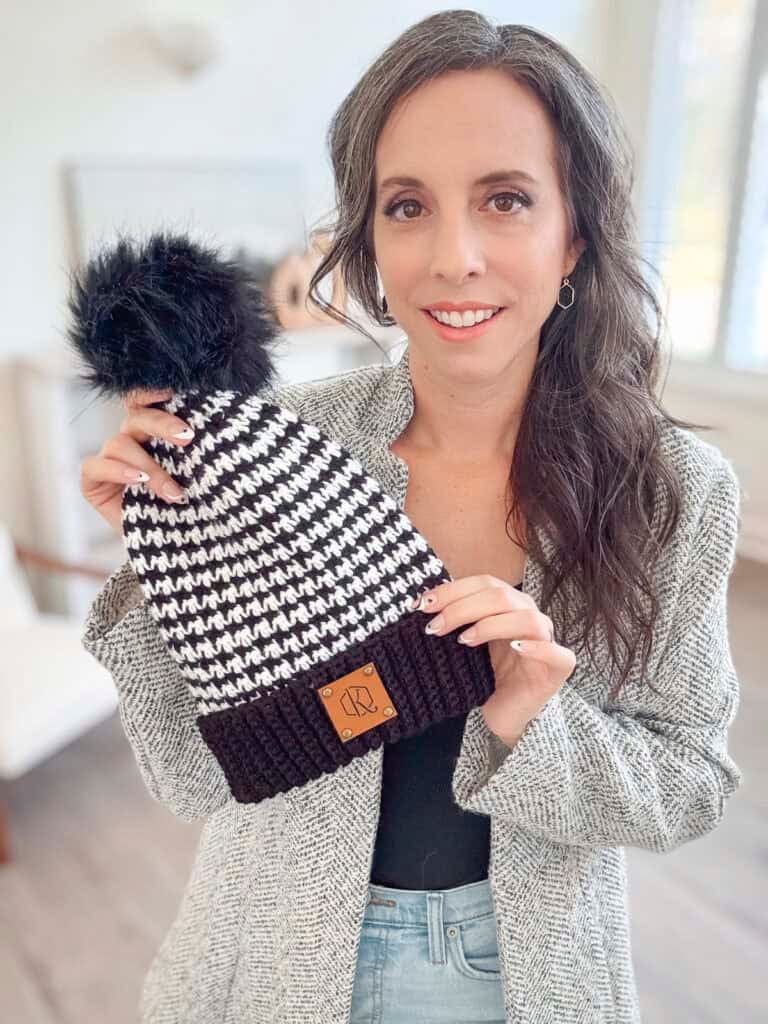 A person holds a black and white houndstooth crochet beanie with a pom-pom, wearing a gray textured coat and standing in a room with natural light.