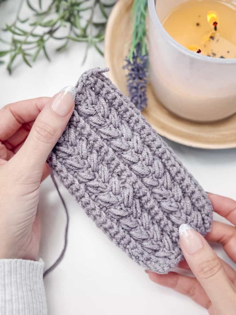 Gentle hands delicately work on a piece of gray crochet, artfully crafting the wheat stitch next to a softly glowing candle, with a lush plant adding a touch of nature in the background.