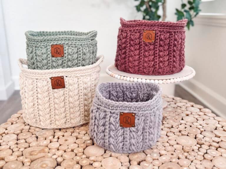 Four knitted baskets in gray, cream, green, and red, each with a leather patch, are placed on wooden surfaces.