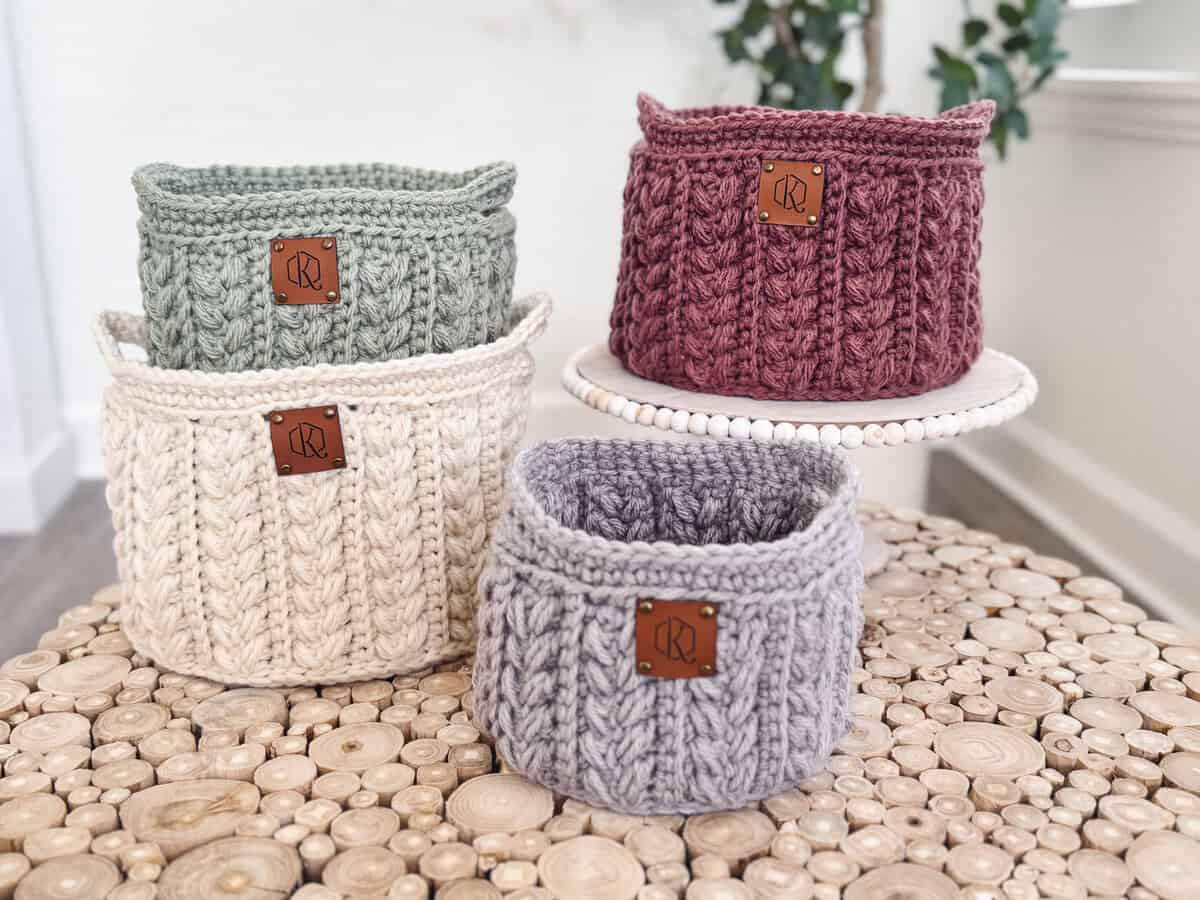 Four knitted baskets in gray, cream, green, and red, each with a leather patch, are placed on wooden surfaces.
